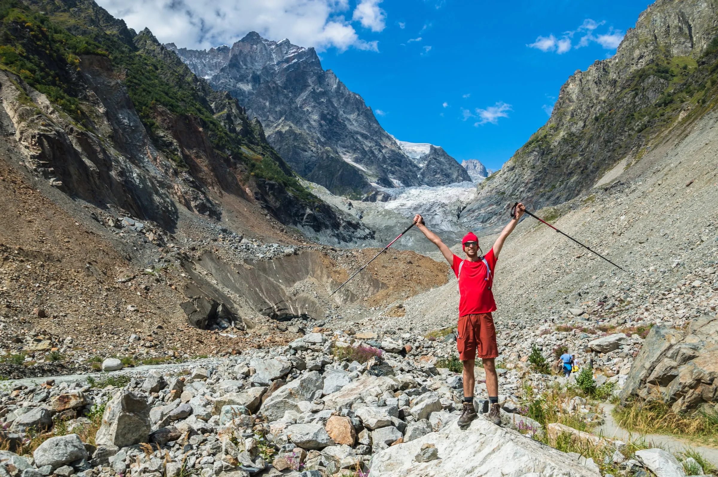 Konrad with arms raised on a rocky glacier trail below snowy peaks in the Caucasus